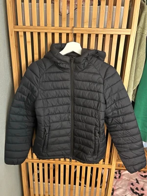 Svart pufferjacka med huva - Svart pufferjacka med huva och dragkedja framtill. Jackan har quiltad design och två fickor med dragkedja. Perfekt för kyligare dagar och enkel att matcha med olika outfits.