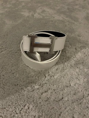 Vändbart vitt och svart bälte med guld H-spänne - Snyggt vändbart bälte i vitt och svart med ett stort H-format metallspänne. Bältet har stilren design och passar perfekt till jeans eller kostymbyxor. Materialet är slätt och ger en exklusiv känsla.