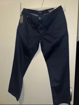 Mörkblå chinos från Replay - Snygga mörkblå chinos från Replay med klassisk rak passform och diskreta fickor både fram och bak. Tillverkade i ett mjukt och bekvämt bomullsmaterial som passar perfekt till en avslappnad men stilren look. Bälteshällor och knappgylf för enkel styling.