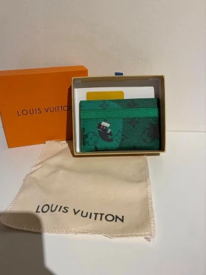 Grönt vuitton kortfodral - Snyggt grönt kortfodral  med klassiskt monogrammönster och en liten detalj med en tecknad figur. Tillverkad i tåligt läder och har flera fack för kort. Kommer med original ask och dustbag.