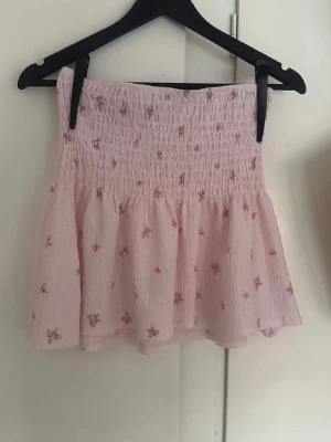 Ljusrosa blommig kortkjol - Supersöt ljusrosa kortkjol med små blommor och smock i midjan. Kjolen har en volangig nederdel och är gjord i ett lätt, luftigt tyg som passar perfekt till varma dagar. Perfekt för dig som gillar romantisk och feminin stil.