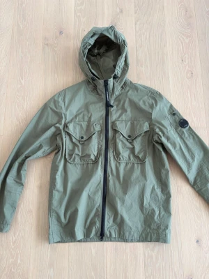Olivgrön vindjacka från C.P. Company - Snygg olivgrön vindjacka från C.P. Company med huva, två stora bröstfickor med lock och knapp samt ikonisk linsdetalj på ärmen. Jackan har dragkedja framtill och är tillverkad i lätt, slitstarkt material som passar perfekt för blåsiga dagar.