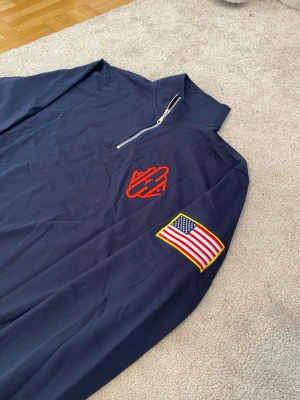 Jeffrey Epstein Bomull Halfzip - Snygg marinblå långärmad tröja, samma som Jeffrey Epstein med halfzip och ribbade muddar. Tröjan har ett rött Carhartt-broderi på bröstet och en amerikansk flagga på ärmen. Perfekt för dig som gillar streetwear och vill ha något unikt.