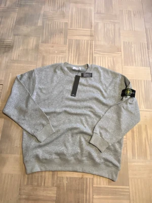 Grå sweatshirt från Stone Island - Snygg grå sweatshirt från Stone Island med klassisk rund hals och ribbade muddar. Ikonisk svart patch med gul och grön logotyp på vänster ärm. Perfekt för dig som gillar streetwear och enkel stil.