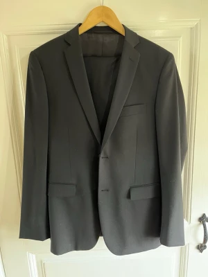 Svart kostym från Dressmann - Stilren svart kostym från Dressmann. Klassisk design med två knappar framtill, fickor med lock och bröstficka. Perfekt för dig som vill ha en snygg och enkel look. Materialet känns mjukt och följsamt. Kavajen storlek 48 regular och byxorna 34/32