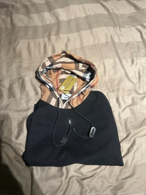 Burrbery hoodie - Snygg svart hoodie från Burberry med klassiskt beige, svart och röd rutmönster på huvan. Hoodien har dragsko i huvan och en stor magficka framtill. Perfekt för dig som vill ha en stilren men ändå unik look. Materialet känns mjukt och bekvämt.