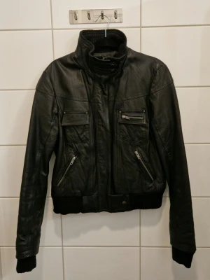 Svart skinnjacka med hög krage - Säljer en svart skinnjacka med hög ribbad krage och snygga detaljer. Jackan har fyra fickor framtill, varav två med dragkedja och två med lock. Ribbstickade muddar vid ärmslut och nederkant. Äkta läder, boxig modell. 