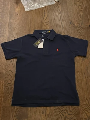 Marinblå pikétröja från Ralph Lauren - Säljer en klassisk marinblå pikétröja från Ralph Lauren i modellen Classic Fit. Tröjan har korta ärmar, krage och två knappar framtill samt den ikoniska röda logotypen broderad på bröstet. Perfekt till jeans eller shorts för en clean och stilren look. Passar M men även S