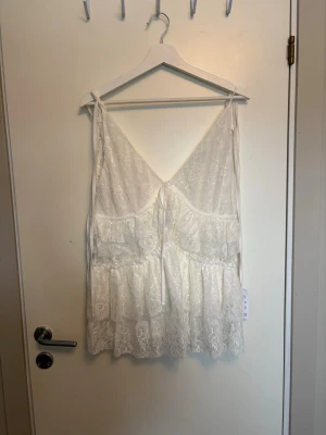 Vit spetslinnetopp från ASOS - Romantisk vit linnetopp från ASOS med tunna axelband, v-ringning och flera lager volanger i spets. Toppen har knyt på axlarna och är perfekt för sommarens alla tillfällen. Söt och luftig design med fina spetsdetaljer.