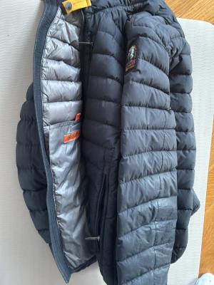 Svart dunjacka från Parajumpers - Snygg svart dunjacka från Parajumpers med quiltad design och gul detalj vid kragen. Jackan har grått innerfoder, praktiska innerfickor och patch med logga på bröstet. Perfekt för kalla dagar och riktigt lätt att bära.