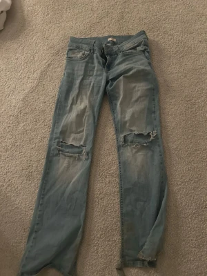 Ljusblå  jeans från Nelly - Säljer ett par ljusblå jeans från Nelly med bootcut ben och stora slitningar över knäna. Jeansen har klassisk femficksmodell, dubbla knappar i midjan och är tillverkade i denim. Perfekta för en avslappnad och trendig look. de är lite slitna vid fötterna❤️