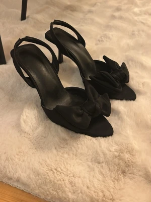 Svarta klackar med rosett - Säljer ett par eleganta svarta klackar med öppen tå och stor dekorativ rosett framtill. Skorna har smal klack och tunna remmar runt hälen för en stilren look. Väldrigt sköna och kanpt använd!