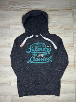 Mörkgrå Superdry hoodie med tryck - Snygg mörkgrå hoodie från Superdry med stort turkost och vitt tryck på bröstet där det står 'Superdry JPN Classics'🙌🏻Hoodien är i fint skick och passar Xs-S🤩 hör av er vid funderingar samt Snabbaffärer🤗