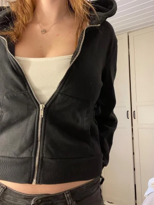 Svart zip hoodie med huva - Säljer en svart hoodie med dragkedja och huva. Modellen är croppad och har breda muddar vid ärmslut och nederkant. Hoodien har fickor framtill och är tillverkad i mjukt sweatshirtmaterial, perfekt för en chill och avslappnad stil.