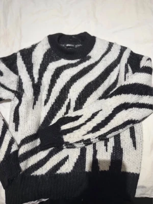 Stickad tröja med zebramönster - Säljer en stickad tröja med svartvitt zebramönster och rund hals. Tröjan har ribbstickade muddar vid ärmslut och nederkant. Perfekt för dig som vill sticka ut med ett djurinspirerat plagg i garderoben.