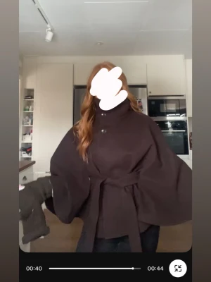 Mörkbrun cape-kappa med bälte - Snygg mörkbrun cape-kappa med hög krage och knäppning framtill. Kappan har vida ärmar och ett matchande knytbälte i midjan som ger en cool siluett. Perfekt för dig som vill ha en trendig och unik look. Materialet ser ut att vara ullblandning.