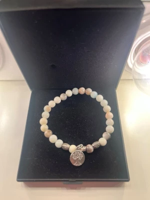 Pärlarmband  - Snyggt armband med runda pärlor i beige och vita toner. Armbandet har en silverfärgad berlock med ett trädmotiv samt små dekorativa metallpärlor. Perfekt accessoar för dig som gillar stilrena detaljer och naturinspirerade smycken.