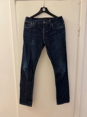 Mörkblå jeans från Nudie Jeans W-32 L-34 - Snygga mörkblå jeans från Nudie Jeans med klassisk femficksdesign och raka ben. Jeansen har orangea kontrastsömmar och är tillverkade i ett slitstarkt denimtyg. Perfekta för en stilren och avslappnad look.