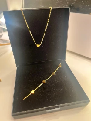 Guldigt halsband med marchande armband - Snyggt halsband i guldfärgad metall med en liten hjärtformad detalj i mitten. Kedjan är tunn och elegant, perfekt för att ge en subtil touch till din outfit. Kommer i en svart ask och har en modern, minimalistisk design.