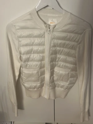 Vit quiltad cardiganjacka från Gina Tricot - fräsch vit cardiganjacka från Gina Tricot med quiltade partier pure fronten, dragkedja och två fickor. Jackan har ribbade i muddar och nederkant samt är tillverkad i en what-deal av stickat och vadderat material för en modern look.