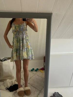 Blommig sommarklänning med volanger - Söt blommig klänning i pastellfärger med volanger och smala axelband. Klänningen har en luftig passform och är perfekt för varma dagar. Mjukt, lätt material som känns skönt mot huden. Färgerna går i gult, grönt, blått och lila.
