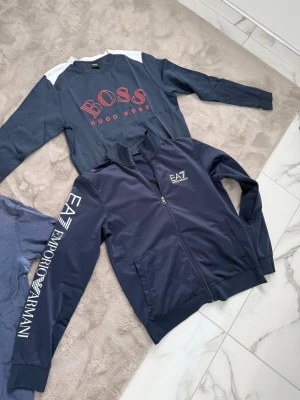 Marinblå hoodie från Emporio Armani - Snygg marinblå hoodie från Emporio Armani med dragkedja framtill och vit logga på bröstet samt text på ärmen. Klassisk passform med ribbade muddar och två fickor. Perfekt för en stilren och sportig look.