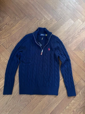 Mörkblå kabelstickad tröja Polo Ralph Lauren - Mörkblå kabelstickad tröja från Polo Ralph Lauren med halv dragkedja och hög krage. Tröjan har det klassiska röda Polo-logot på bröstet och ribbade muddar. Perfekt för lager på lager och snygg till jeans.