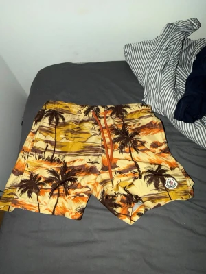 Moncler badbyxor med palmtryck - Snygga badbyxor från Moncler med tropiskt palmtryck i gula, orangea och bruna toner. Klassisk modell med resår och dragsko i midjan samt Moncler-logga på ena benet. Perfekta för stranden eller poolen. Har använts en gång. Erbjud ett annat pris så kanske jag går med på det 