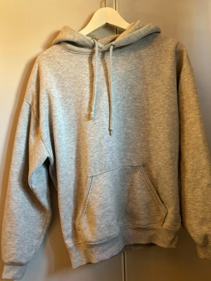 Ljusgrå basic hoodie med ficka - En stilren ljusgrå hoodie med huva och dragsnören. Hoodien har en stor magficka och ribbade muddar vid ärmslut och nederkant. Perfekt för en avslappnad look och enkel att matcha med andra plagg.