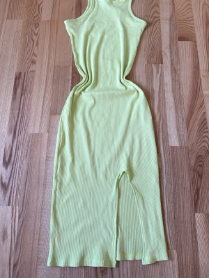 Ljusgul ribbad långklänning - Det är lite öppen vid benen men går att sy ihop det som har gått lite sönder ni kan se på bilden och baksidan uppe på ryggen så har den fått röd fläck syns inte så mycket om du har utsläpp hår, Snygg ljusgul långklänning i ribbat material med ärmlös design och rund halsringning. Klänningen har en slits framtill som ger en cool touch. Perfekt för dig som gillar en enkel men trendig stil.