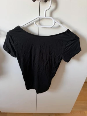 Svart topp med rosett i ryggen - Svart kortärmad topp med bred urringning och en snygg satinrosett i ryggen. Toppens material känns mjukt och stretchigt, perfekt för en trendig look. Passformen är normal och toppen är enkel att matcha med olika outfits.