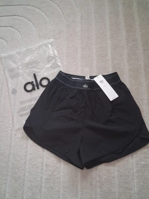 Svarta träningsshorts från Alo Yoga - Snygga svarta träningsshorts från Alo med elastisk midja och vit kontrastsöm. Lätta och luftiga i syntetmaterial, perfekta för aktiva dagar. Klassisk sportig look med diskret logga både fram och bak.