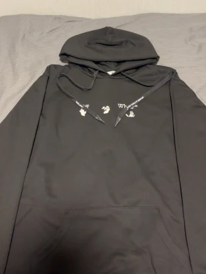Svart hoodie med tryck och stor ryggdesign - Svart hoodie från Who Decides War med vit text och små symboler på bröstet samt en stor, unik dödskalle- och korsben-illustration i lila och vitt på ryggen. Hoodien har huva med dragsko och en klassisk magficka. Perfekt för dig som gillar streetwear och coola prints.