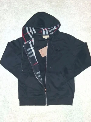 Svart hoodie från Burberry - Svart hoodie med dragkedja från Burberry. Klassisk huva med det ikoniska rutmönstret på insidan och röda detaljer. Mjukt material och två fickor framtill. Perfekt för en stilren och avslappnad look. Skriv privat vid frågor.