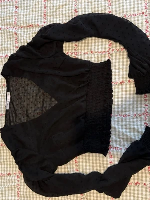 Svart omlottopp med prickig struktur - Trendig svart omlottopp från NA-KD med långärmade puffärmar och smockad midja. Tunt, lätt material med diskret prickig struktur.