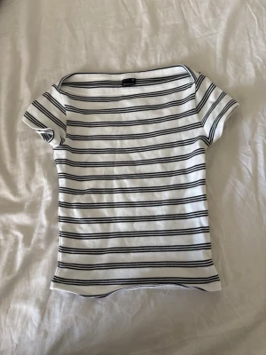 Randig topp från Gina Tricot - Vit ribbad topp från Gina Tricot med svarta horisontella ränder. T-shirten har korta ärmar och en klassisk, något bredare rund halsringning. Perfekt för en avslappnad och stilren look.