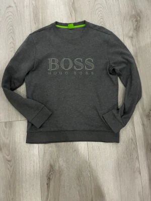 Grå sweatshirt från Hugo Boss - Snygg grå sweatshirt från Hugo Boss med broderad logga på bröstet. Tröjan har rund halsringning, ribbade muddar och är i slim fit-modell. Perfekt för en stilren och avslappnad look.