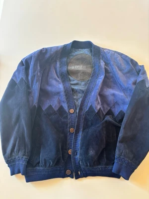 Blå mocka-cardigan från Terras - Snygg blå cardiganjacka i mocka från Terras. Jackan har knäppning med bruna knappar, ribbade muddar och ett coolt geometriskt mönster på fodret. Perfekt för dig som gillar retrostil och unika detaljer.