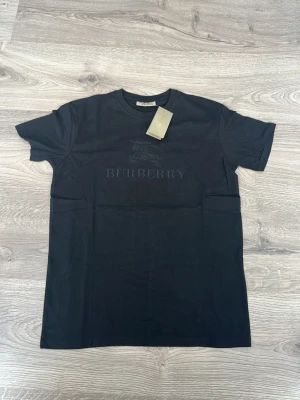 Svart Burberry t-shirt med logga - Snygg svart t-shirt från Burberry med diskret broderad logga och hästmotiv på bröstet. Klassisk passform och rund halsringning. Perfekt för dig som gillar stilrena plagg med exklusiv känsla.