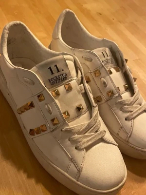 Vita Valentino Rockstud sneakers - vita sneakers från Valentino med guld nitar. de är köpta förra året och andvända 1 gång. Säljer pga att de är för stora för mig. Priset kan diskuteras 