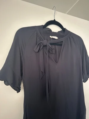 Svart blus med knytband - Snygg svart blus med korta volangärmar och dekorativt knytband vid halsen. Blusen har en lätt v-ringning och är tillverkad i ett mjukt, tunt tyg som faller fint. Perfekt för en trendig och stilren look.