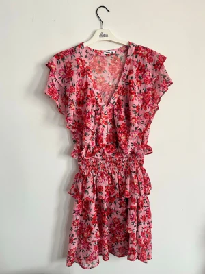 Blommig volangklänning  - Supersöt klänning från Replay i rosa med blommigt mönster i rött och grönt. Klänningen har volangdetaljer, v-ringning och korta ärmar. Perfekt för sommarens alla tillfällen.