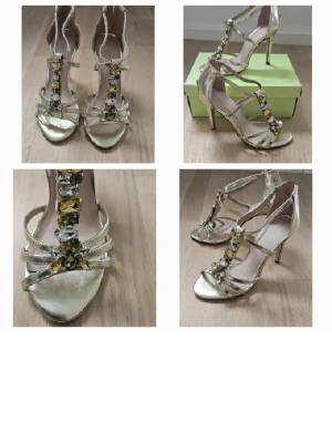 Guldiga sandaler med strassdetaljer - Snygga sandaler i metallic guld med smala remmar och höga klackar. Framtill pryds de av stora glittrande strass i guld och silver. Perfekta för att sticka ut och ge extra bling till din outfit. Öppen tå och dragkedja bak. klackhöjd 10 cm. Använda endast 2 gånger.