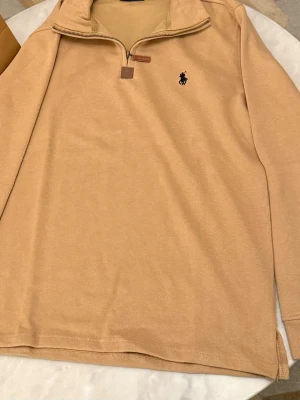 Beige stickad tröja Polo Ralph Lauren - Snygg beige stickad tröja från Polo Ralph Lauren med halv dragkedja och klassisk broderad logga på bröstet. Tröjan har hög krage och ribbade muddar, perfekt för lager-på-lager. Materialet känns mjukt och bekvämt.