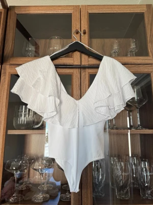 Vit volangbody Zara - Supersnygg vit body med djup v-ringning och stora volangdetaljer över axlarna, figurnära passform. Perfekt till en kjol eller ett par jeans nu till sommaren. Bodyn är köpt ifrån Zara förra året för 300kr. Aldrig använt då den tyvärr aldrig passade!🫶🏼