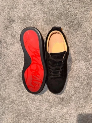 Svarta Christian louboutin skor - Säljer ett par svarta sneakers från Christian Louboutin i mjuk mocka med klassisk röd sula och signaturdetaljer undertill. Skorna har lågt skaft, svarta skosnören och en stilren design som sticker ut med den ikoniska röda undersidan.