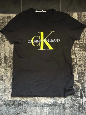 Svart Calvin Klein t-shirt med tryck - Svart t-shirt från Calvin Klein Jeans med stort gult CK-tryck och vit text på bröstet. Klassisk rund hals och korta ärmar. Perfekt för en stilren och trendig look.