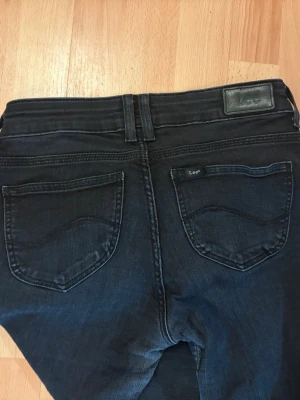 Mörkblå slim fit jeans från Lee - Snygga mörkblå jeans från Lee med slim fit-modell och klassiska fem fickor. Jeansen har Lee-logga på bakfickan och läderpatch i midjan. Perfekta för en trendig och avslappnad stil. Materialet är mjukt denim med lite stretch för extra komfort.