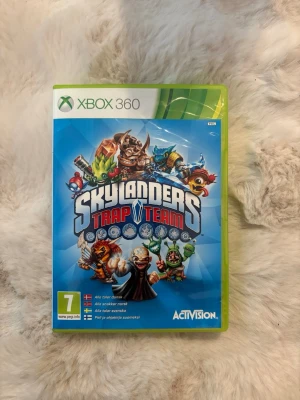 Skylanders Trap Team Xbox 360 spel - Skylanders Trap Team till Xbox 360 är ett actionäventyrsspel där du fångar skurkar och använder dem i spelet. Omslaget är blått med färgglada karaktärer och spelet levereras i ett klassiskt grönt Xbox 360-fodral. Perfekt för dig som gillar fantasy och samlarfigurer.