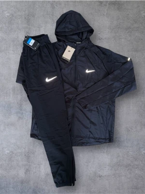 Svart träningsset från Nike med huva - Säljer ett svart träningsset från Nike med jacka och byxor. Jackan har huva, dragkedja och två fickor samt reflekterande Nike-logga på bröstet. Byxorna har resår i midjan och reflekterande logga. Materialet är lätt och passar perfekt för träning utomhus.